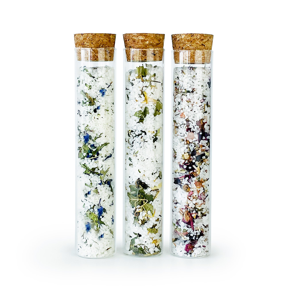 Drei Glasröhrchen mit Korkverschluss, gefüllt mit verschiedenen Badesalzen und getrockneten Blumen.