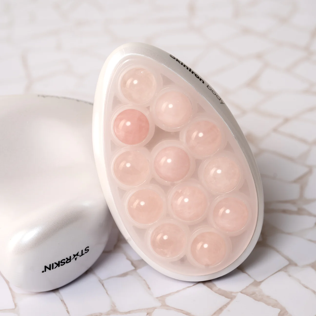 231128_skinironbody2_1296x Ein weißes Hautpflegegerät mit mehreren runden, rosafarbenen Kugeln auf einer Marmoroberfläche.