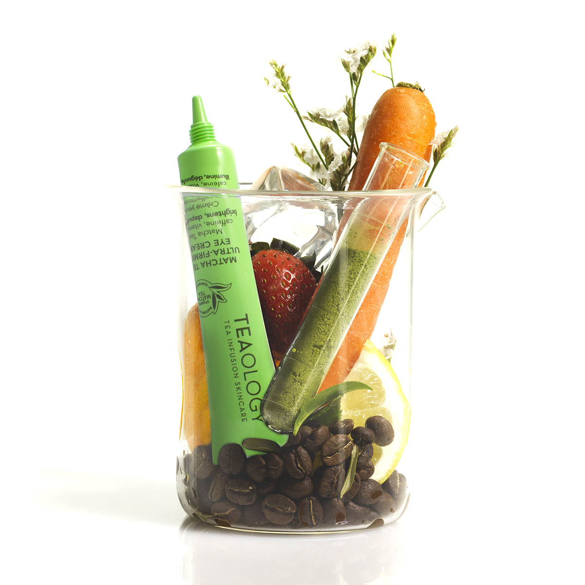 Ein Becherglas mit Kaffeebohnen, Karotte, Erdbeere, Zitrone, Reagenzglas und Tealogy Matcha Augencreme.