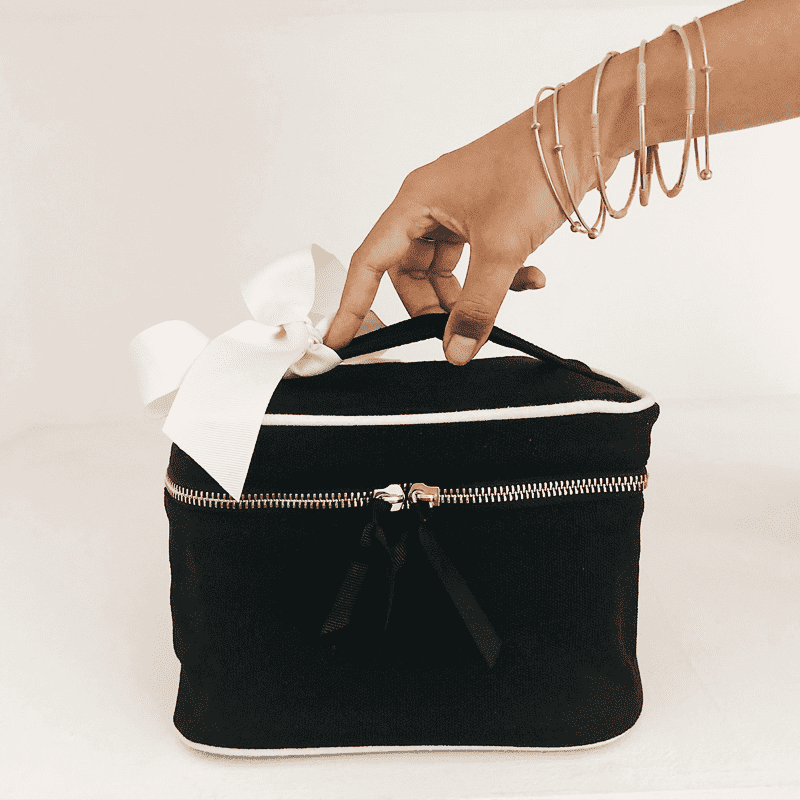 Hand greift nach schwarzer Kosmetiktasche mit weißer Schleife auf weißem Hintergrund.